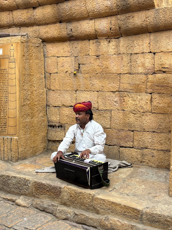 A street musician in Jaisalmer, India playing the harmonium against a historic sandstone wall.